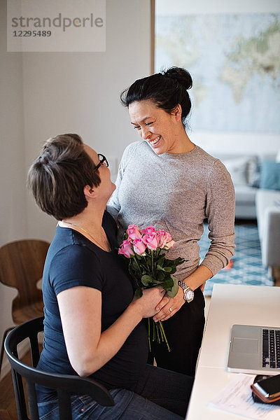 Lesbian couple talking and embracing while holding pink roses at home