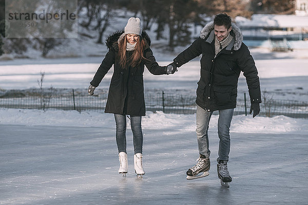 Volle Länge des Mannes  der mit der Frau Händchen hält  während er auf der Eisbahn Schlittschuh läuft.