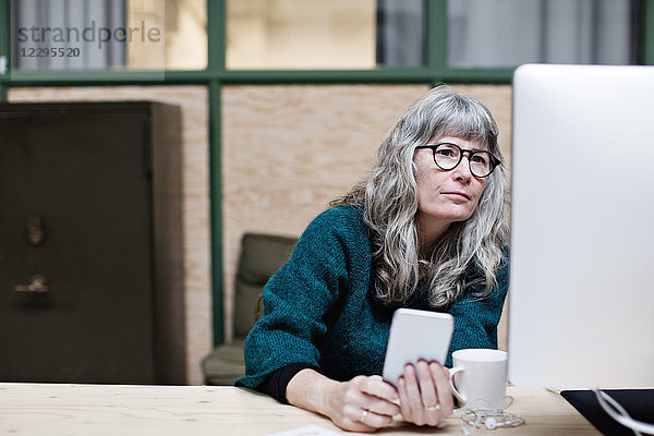 Serious mature businesswoman with mobile phone looking at computer in office