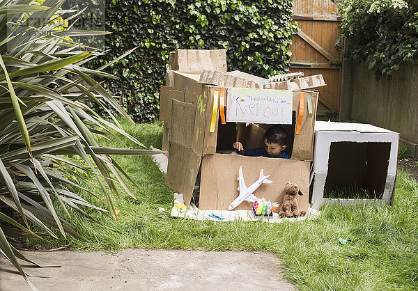 Junge im Pappspielhaus mit Spielzeug im Hinterhof