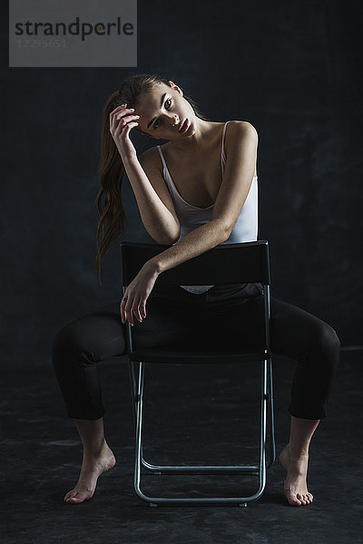 Ganzflächiges Porträt eines jungen Models  das auf einem Stuhl auf schwarzem Hintergrund sitzt.