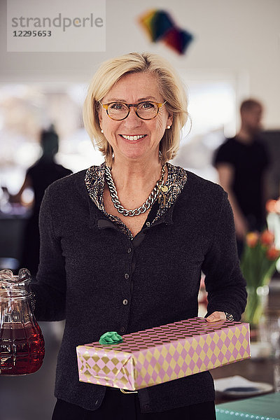 Portrait of smiling senior woman holding gift box and juice while standing at home