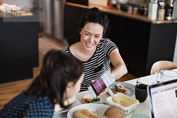 Smiling mother showing mobile phone to daughter while having breakfast at dining table
