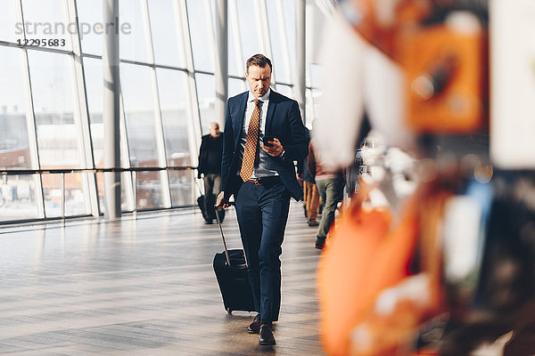 Full length of mature businessman with luggage using mobile phone while walking in airport terminal