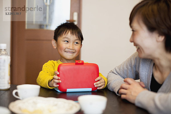 Lächelnder Junge mit Lunchbox und Blick auf die Mutter im Haus