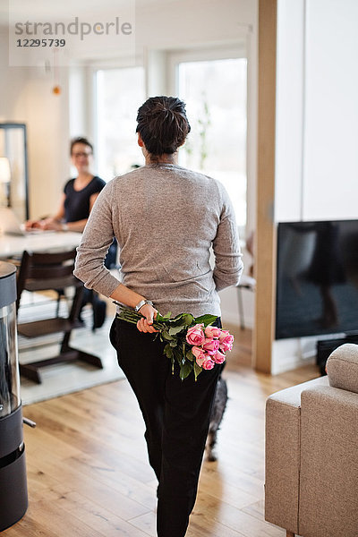 Rear view of woman hiding bouquet while walking towards girlfriend at home