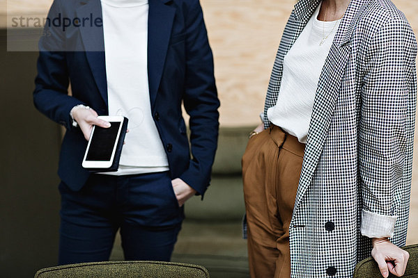 Midsection of businesswoman holding mobile phone while standing with female professional at office