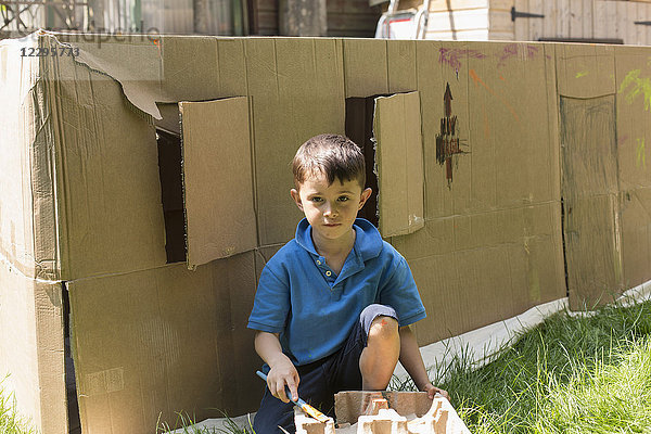 Porträt eines Jungen  der an einem sonnigen Tag gegen ein Pappspielhaus im Hinterhof malt.
