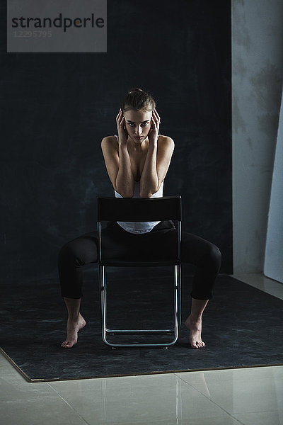 Ganzflächiges Porträt eines jungen Models auf einem Stuhl vor schwarzem Hintergrund.