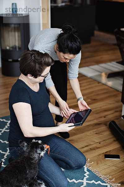 Woman showing digital tablet to girlfriend sitting by dog on carpet in living room
