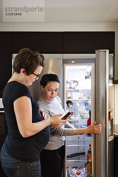 Woman showing mobile phone to girlfriend while standing by open refrigerator in kitchen
