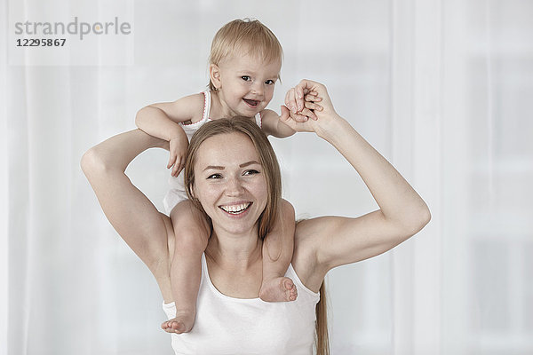 Glückliche Mutter trägt Tochter auf Schultern gegen weißen Vorhang zu Hause