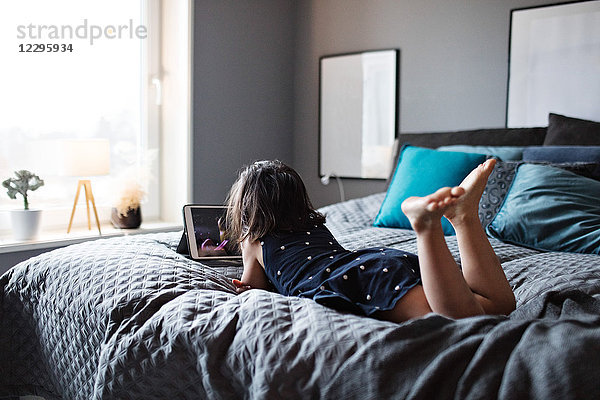 Full length of girl watching digital tablet while lying on bed at home