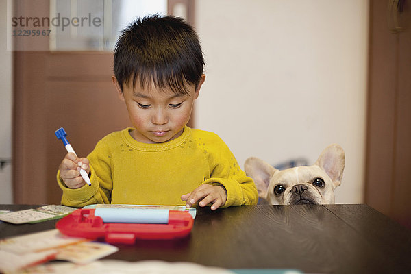 Junge schreibt auf Schiefer  während er am Tisch neben dem Hund steht.