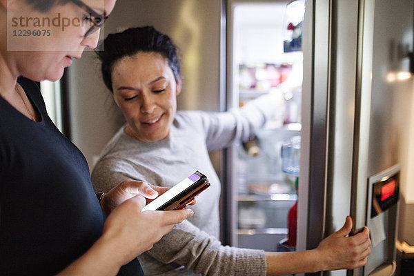 Woman showing mobile phone while girlfriend opening refrigerator in kitchen