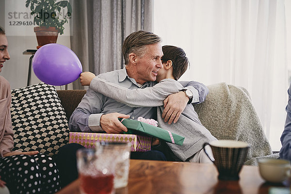 Grandfather holding birthday gift while embracing grandson on sofa at home