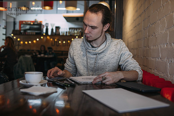 Junger Mann beim Sitzen mit Handy und Buch auf dem Tisch im Cafe