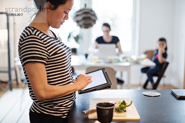 Woman making food while using digital tablet at kitchen with family in background