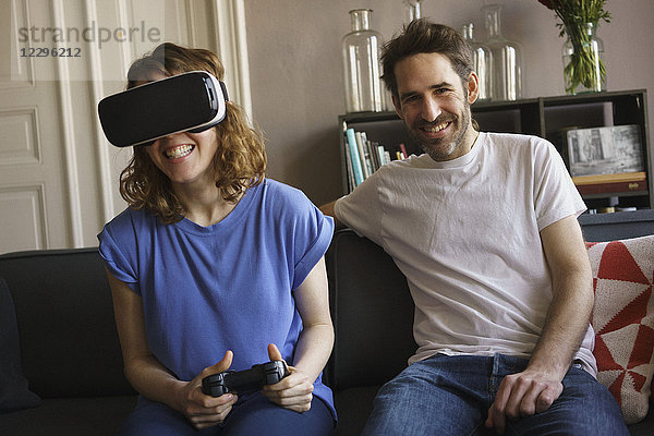 Porträt eines lächelnden Mannes  der von einer Frau auf einem Virtual-Reality-Headset im Wohnzimmer sitzt.