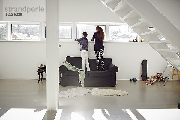 Full length of siblings standing on sofa while looking through window at home