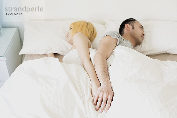 Hochwinkliger Blick auf das Paar  das sich an den Händen hält  während es Rücken an Rücken im Bett schläft.