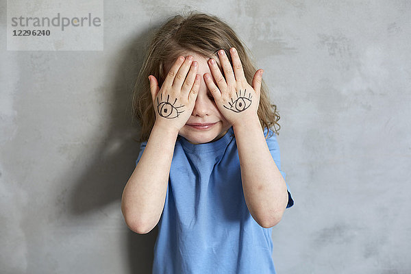 Nahaufnahme eines Mädchens  das die Augen mit bemalten Händen bedeckt  während es an einer grauen Wand steht.