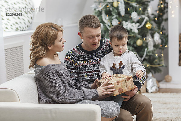 Mann schaut Frau an  die dem Sohn  der zu Hause auf dem Sofa sitzt  zu Weihnachten ein Geschenk macht.