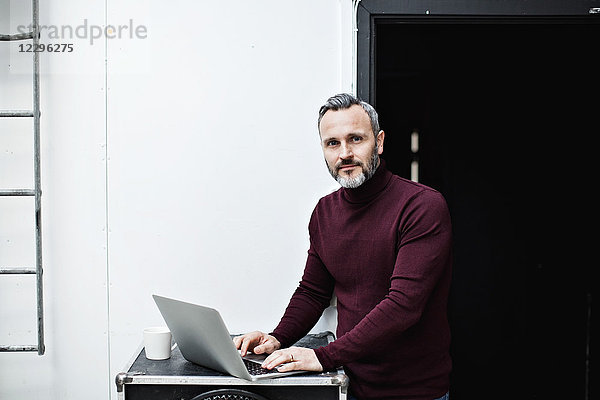 Portrait of mature businessman using laptop while standing by wall at creative office