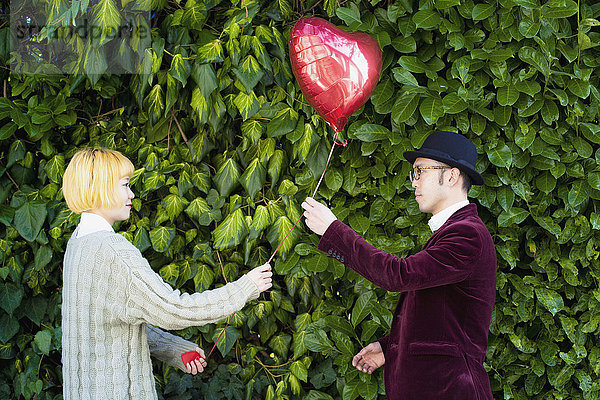Seitenansicht eines jungen Paares  das einen herzförmigen Ballon gegen Pflanzen im Park hält.