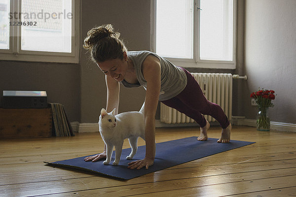 Lächelnde Frau übt Plankenposition über weißer Katze auf Trainingsmatte zu Hause
