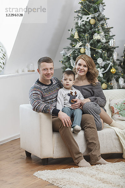 Porträt eines lächelnden Vaters und einer lächelnden Mutter  die mit ihrem Sohn auf dem Sofa am Weihnachtsbaum sitzen.