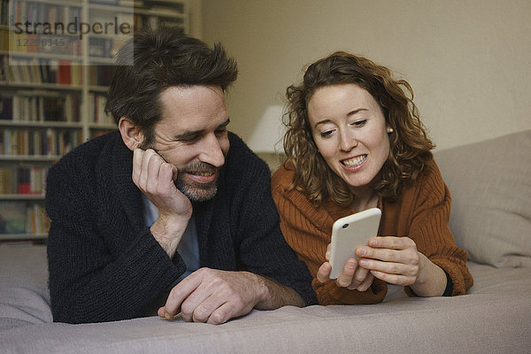 Lächelnde Frau teilt sich das Smartphone mit dem Mann  der zu Hause im Bett liegt.