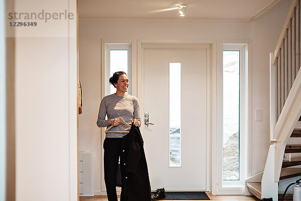 Smiling woman looking away while hanging coat to coathanger against door at home