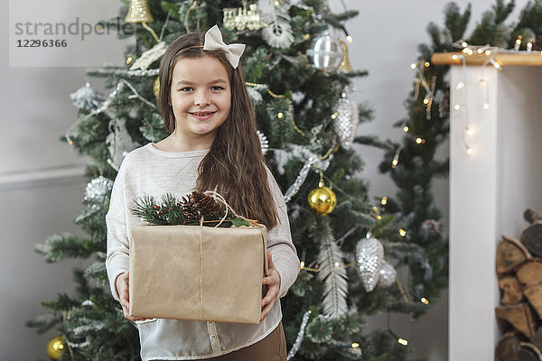 Porträt des lächelnden Mädchens mit Geschenk gegen den Weihnachtsbaum zu Hause