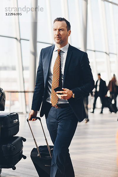 Confident mature businessman with mobile phone and luggage walking in airport