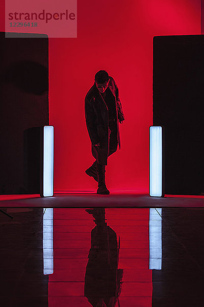 Volle Länge des jungen Mannes inmitten von beleuchteten Lichtern mit Reflektion am Boden im roten Studio