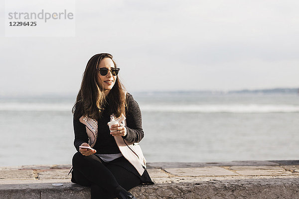 Lächelnde Frau sitzt auf einer Stützmauer mit Smartphone und trinkt gegen das Meer.