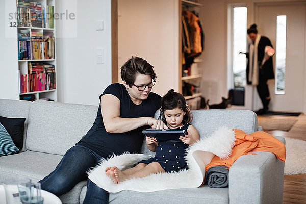 Mother showing digital tablet to daughter while sitting on sofa at home