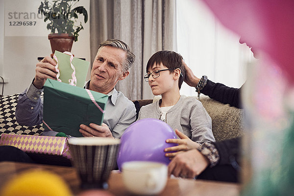 Boy and grandfather looking at greeting card and birthday present while sitting on sofa