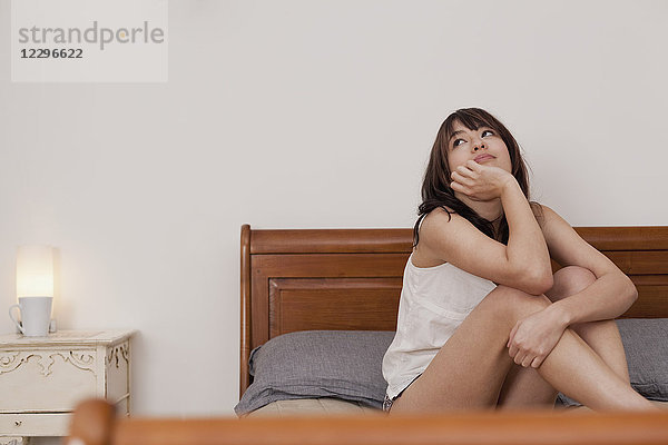 Nachdenkliche junge Frau sitzt zu Hause auf dem Bett gegen die Wand.