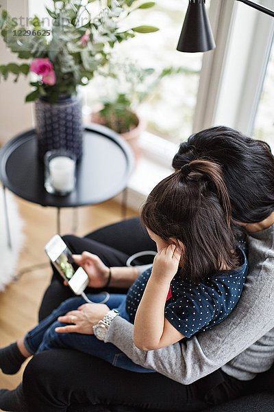 High angle view of mother and daughter wearing headphones while using mobile phone in living room