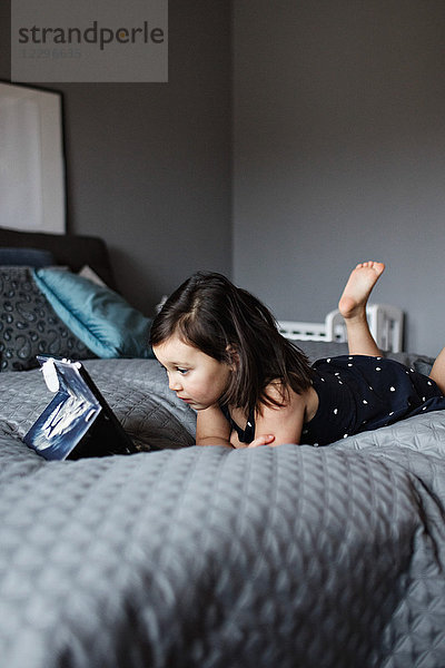 Full length of cute girl watching digital tablet while lying on bed at home