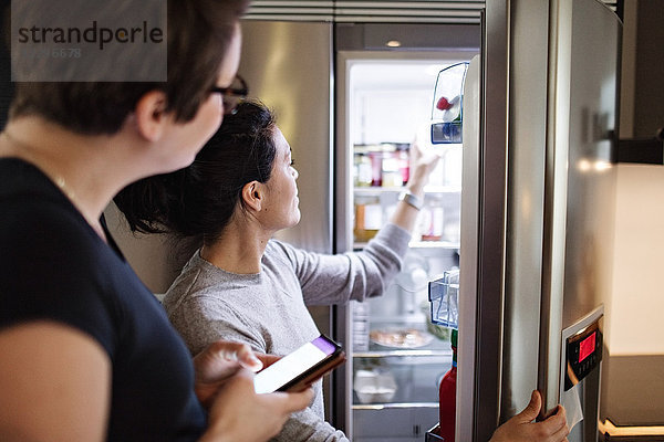 Woman with mobile phone looking at girlfriend opening refrigerator in kitchen