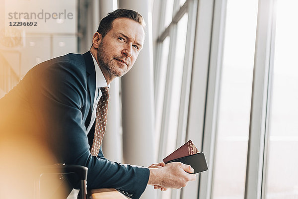 Confident mature businessman with passport and mobile phone leaning on railing at airport