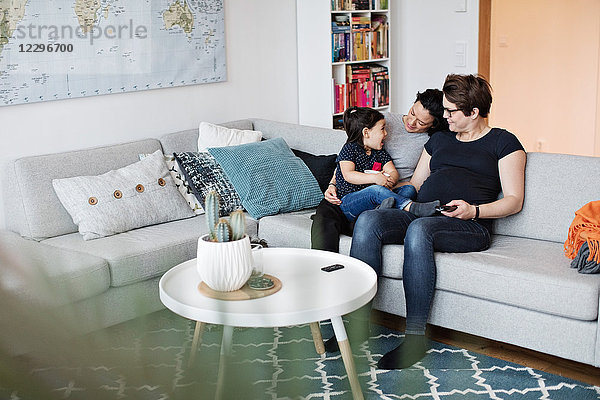 Smiling lesbian couple sitting with daughter on sofa in living room at home