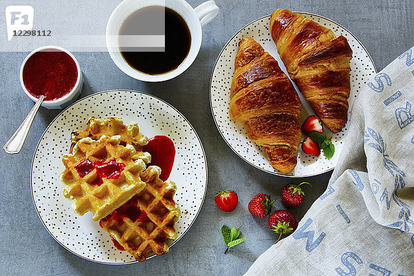 Nahaufnahme des Frühstückstisches mit frisch gebackenen Croissants und Waffeln  serviert mit Kaffee  Erdbeeren und hausgemachter Beerenmarmelade