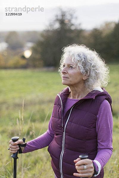 Thoughtful active senior woman hiking with poles in rural field
