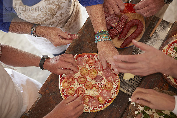 Overhead view senior friends adding tomatoes and meat to fresh pizza
