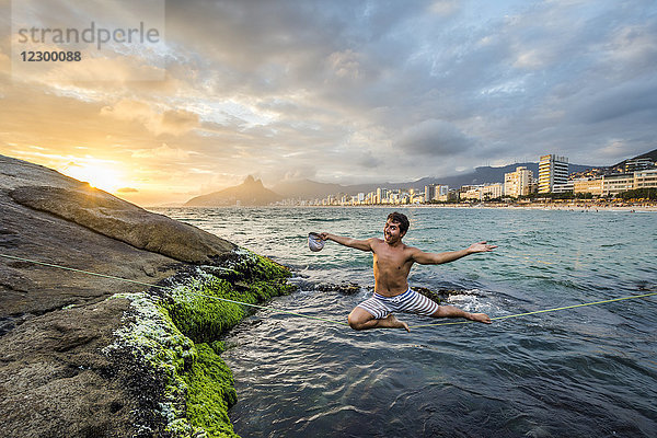 Junger Mann schaut in die Kamera  während er lächelnd auf einer Slackline balanciert  die bei Sonnenuntergang über dem Wasser gespannt ist  Rio de Janeiro  Brasilien