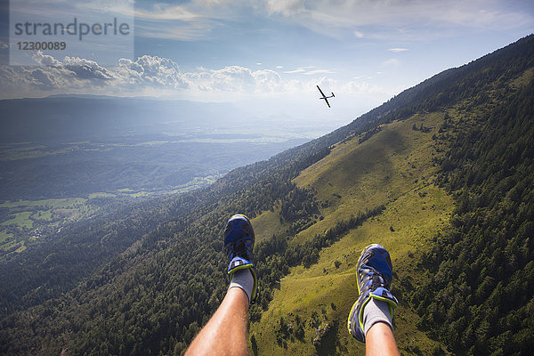 Die Füße eines Gleitschirmfliegers baumeln hoch über den slowenischen Alpen bei einem Gleitschirmflug vom Berg Dobrca bei Begunje und Radovljica in Slowenien. Im Hintergrund ein Segelflugzeug.
